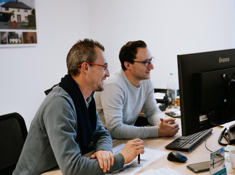 Deux hommes souriants travaillant ensemble devant un ordinateur. Documents et plans d’architecte sur le bureau.
