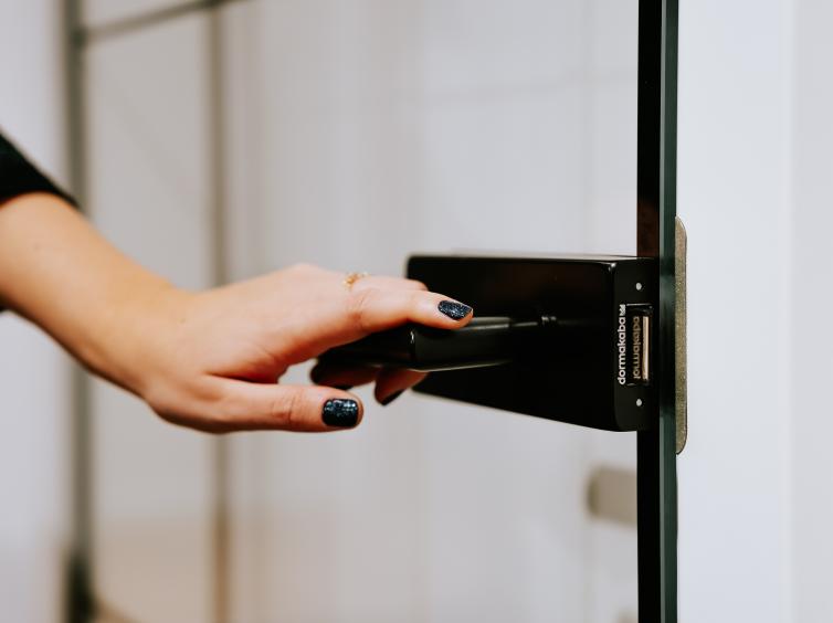 Une main ouvrant une porte en verre avec une poignée noire moderne. Ongles vernis et bague fine en détail.