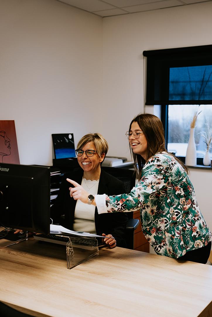 Deux femmes souriantes dans un bureau, travaillant ensemble sur un ordinateur. L’une est assise, vêtue d’un blazer noir, et l’autre, debout, porte une veste colorée et pointe l’écran.