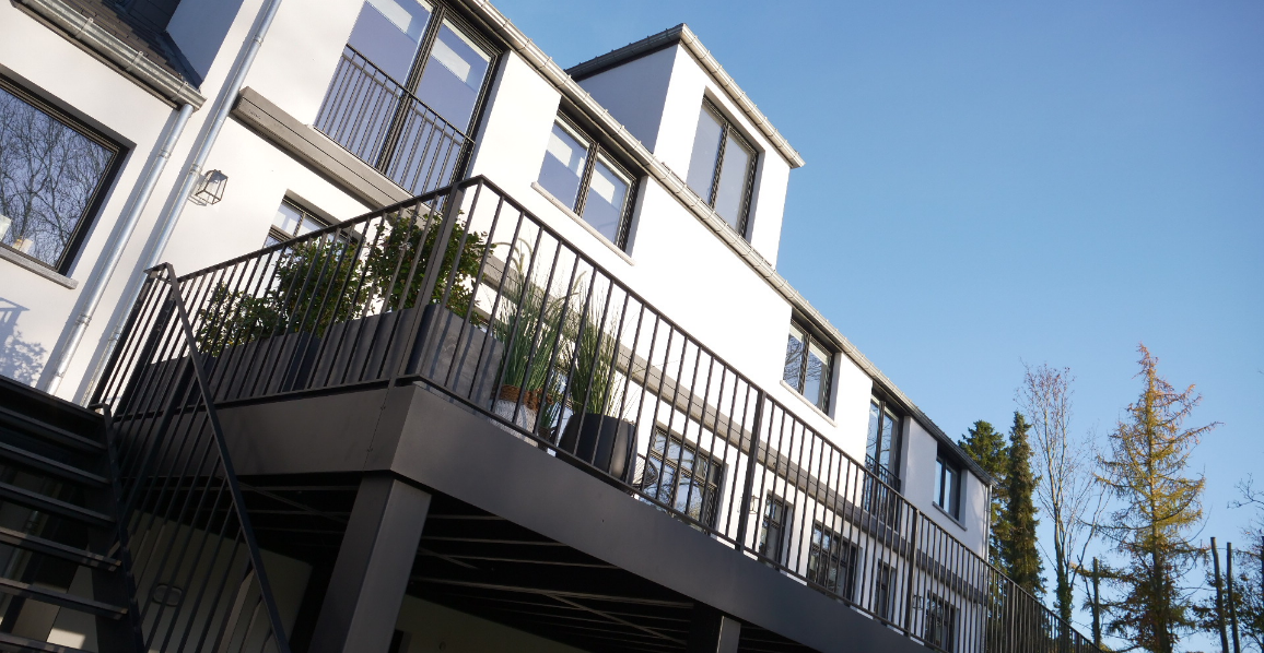Maison moderne à façade blanche avec de grandes fenêtres et un toit en pente. Une terrasse surélevée en métal noir avec garde-corps accueille des plantes en pots.