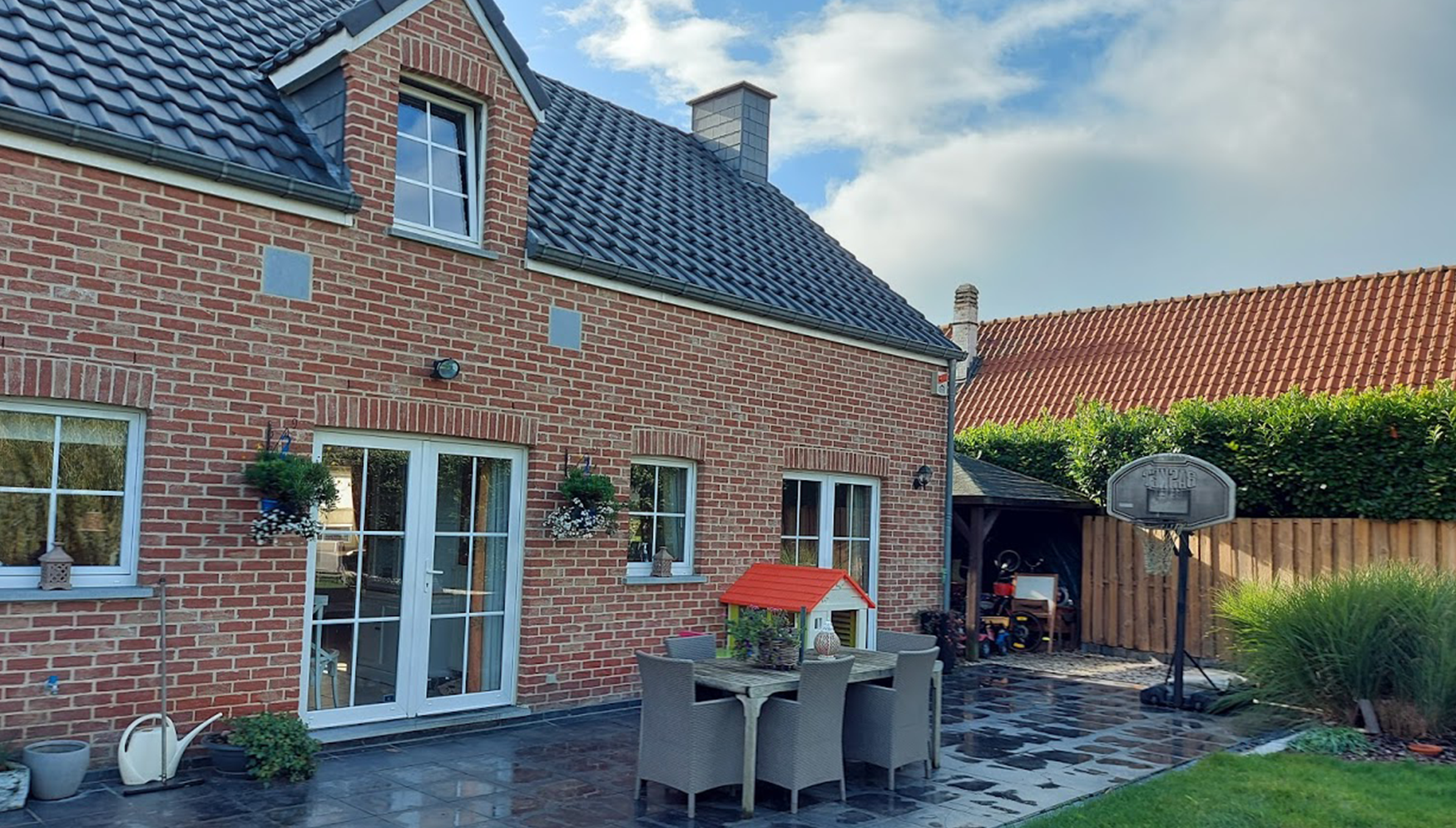 Maison en briques rouges avec toit en tuiles noires et grandes fenêtres blanches. Terrasse carrelée avec salon de jardin et niche rouge pour chien. Panier de basket et abri de rangement en arrière-plan.