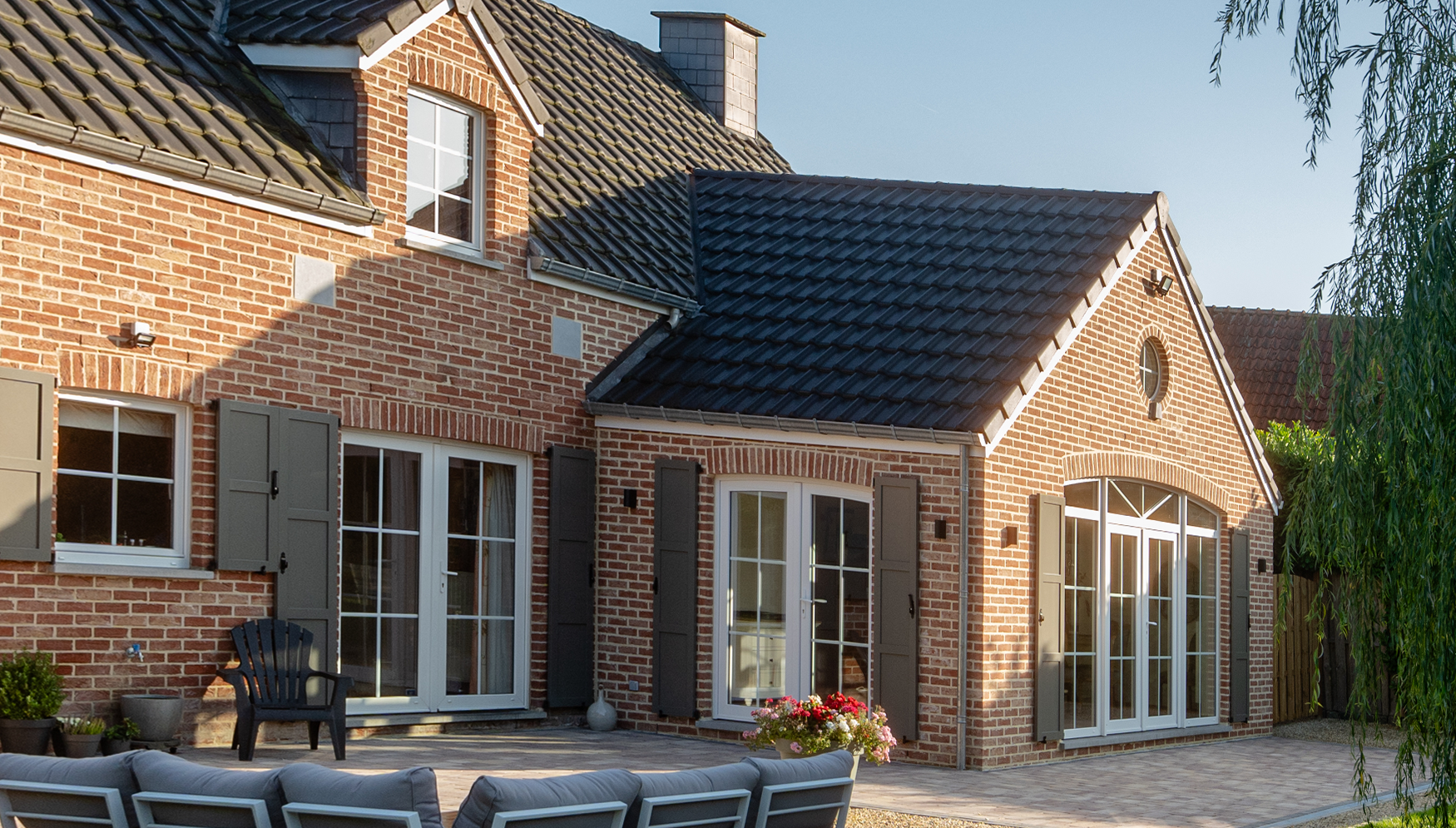 Maison en briques rouges avec toit en tuiles noires, volets gris et grandes portes-fenêtres blanches. Une terrasse en pierre avec mobilier d'extérieur complète l’ensemble.